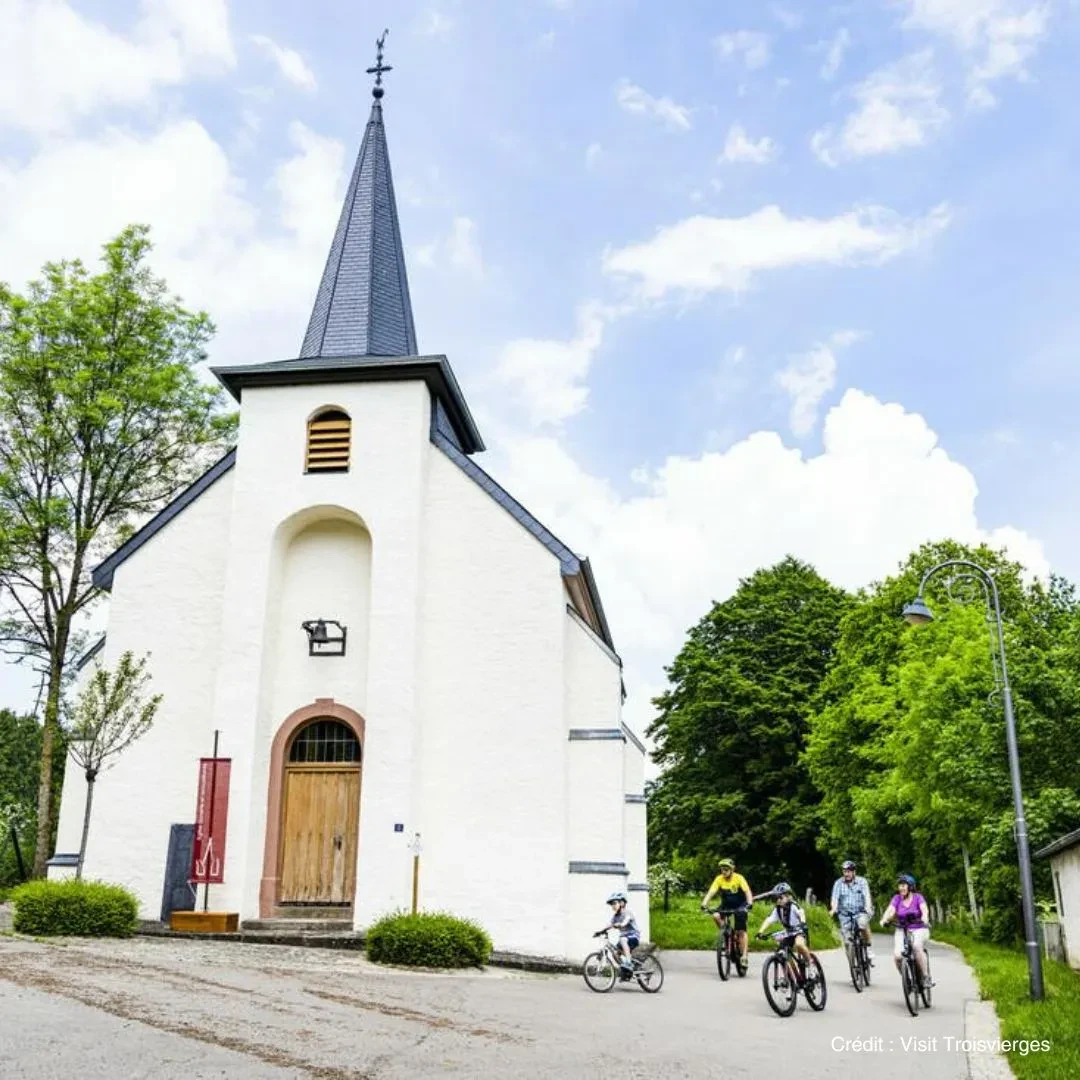 Église blanche avec cyclistes devant arbres - Séjour Bien-être Luxembourg - Anatura Luxembourg